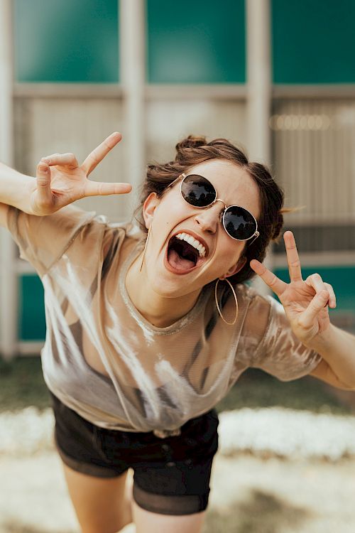 A person wearing sunglasses and making peace signs with both hands, smiling widely. They appear to be enjoying themselves in a casual setting.