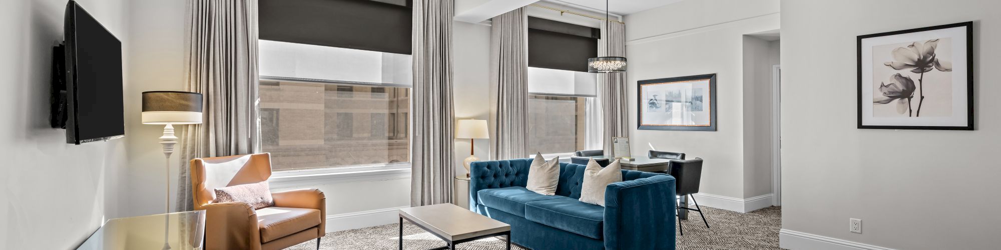 A modern living room with a blue sofa, armchair, TV, and wall art; features carpet flooring and a ceiling light fixture.