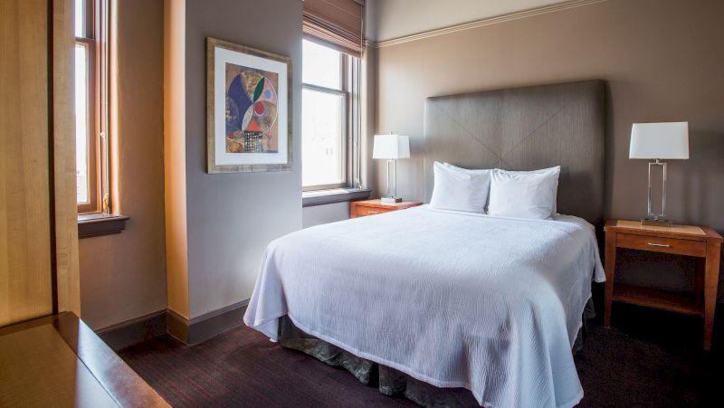 A neatly made bed with white linens and pillows, two wooden nightstands with lamps, and a piece of abstract artwork on the wall in a hotel room with a window.
