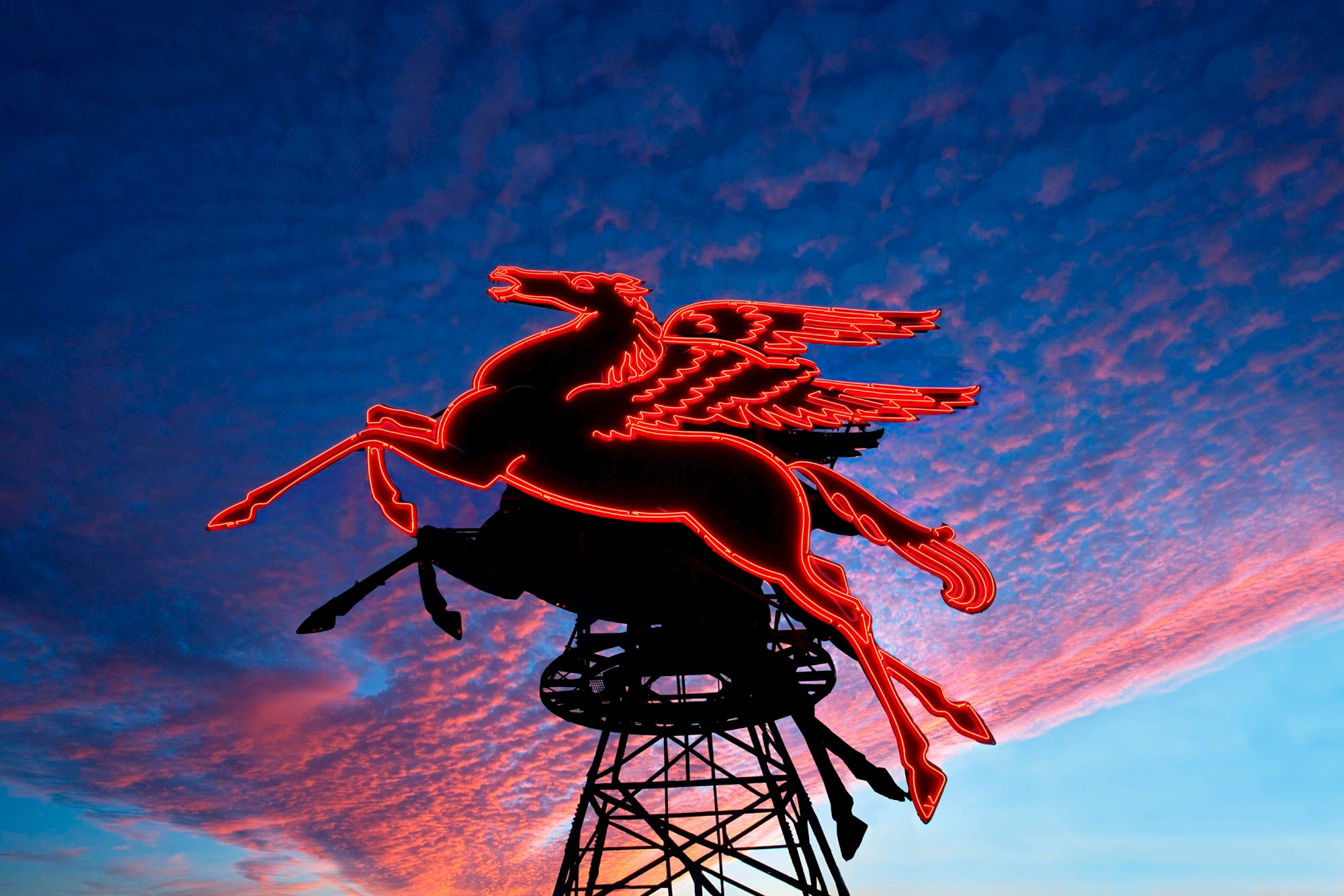 A glowing red neon Pegasus sign sits atop a metal structure against a dramatic, colorful sky at dusk.