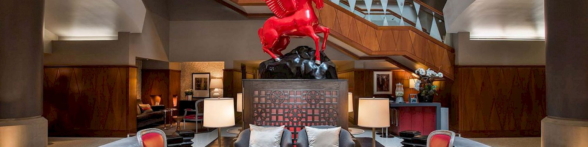 A lobby with seating arrangements, a red Pegasus statue centerpiece, and a grand staircase in the background.