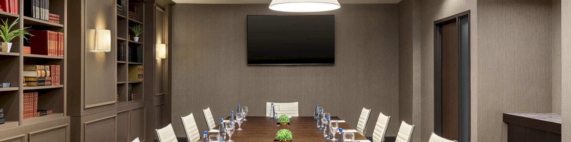 A modern conference room with a long table, chairs, a screen on the wall, bookshelves, and notepads with water glasses on the table.