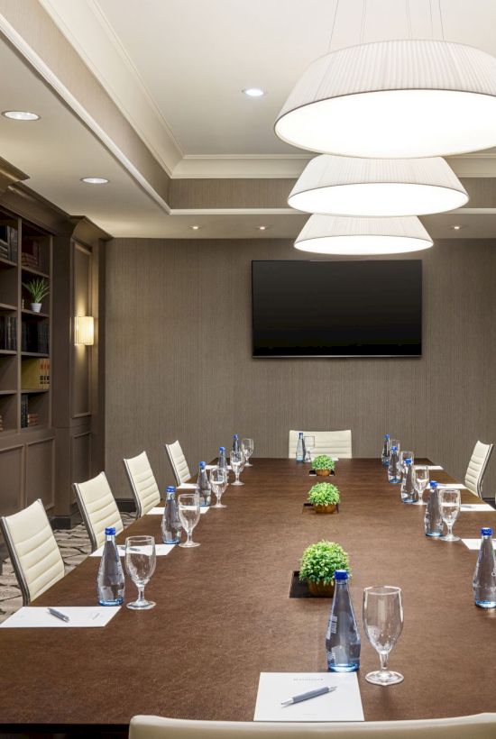 A sleek conference room with a long table, surrounded by white chairs, plants, water glasses, notepads, and a screen on the wall.