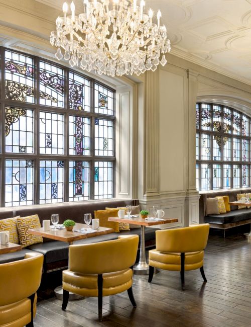 The image shows a luxurious dining area with chandeliers, stained glass windows, and yellow chairs around wooden tables elegantly set.