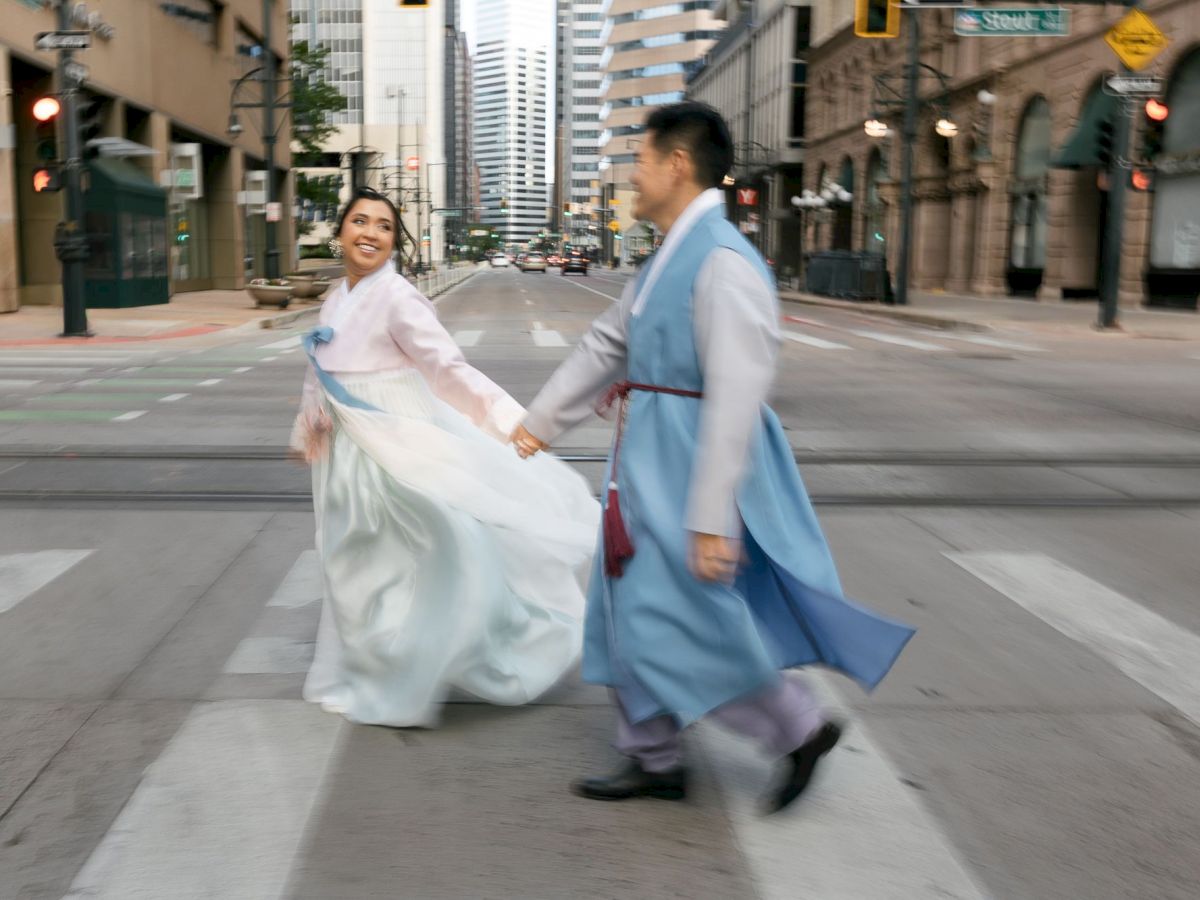 A couple in traditional attire walks hand in hand across a city street, surrounded by tall buildings and urban scenery.
