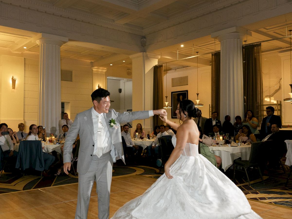 A couple is dancing in a large, elegant hall with guests seated around tables, watching them.