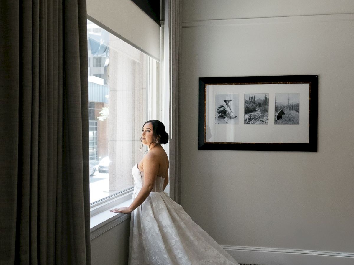 A person in a wedding dress stands by a window, gazing outside. The room has a carpeted floor and black-and-white photographs on the wall.
