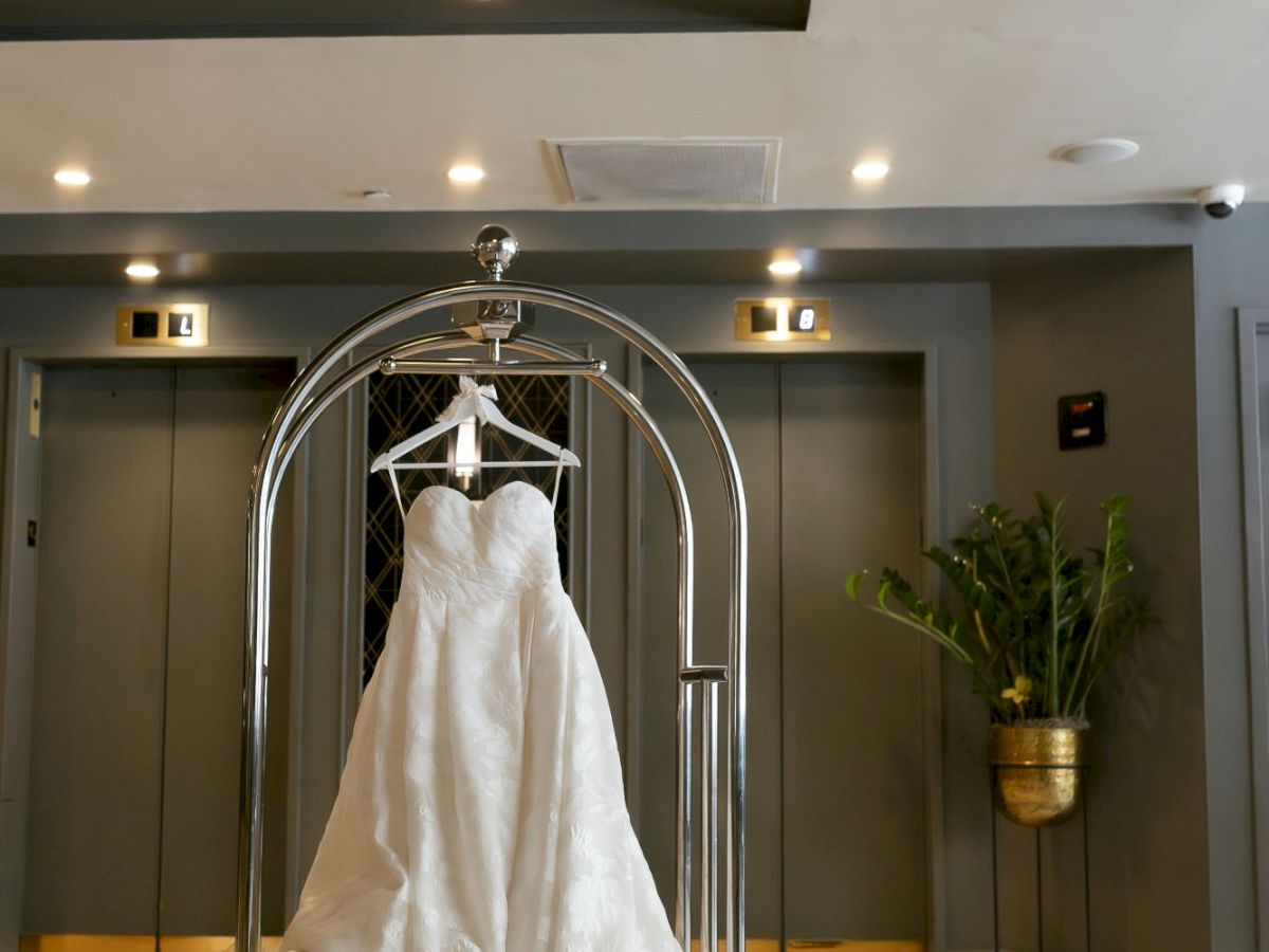 A white wedding dress is displayed on a luggage cart in a modern hallway with elevators and plants.