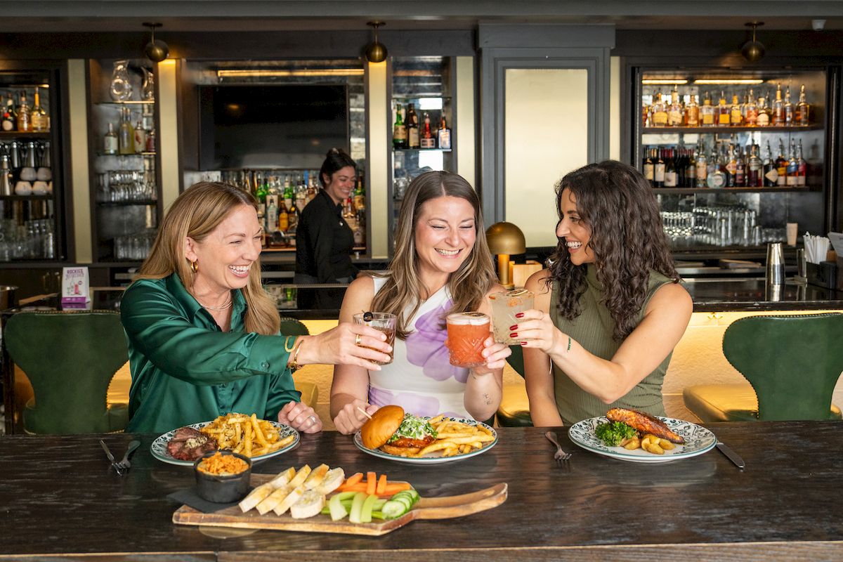 Three women are toasting drinks over plates of food in a stylish restaurant or bar setting.