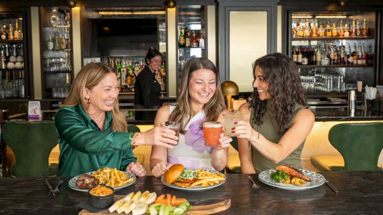 Three women are toasting drinks over plates of food in a stylish restaurant or bar setting.