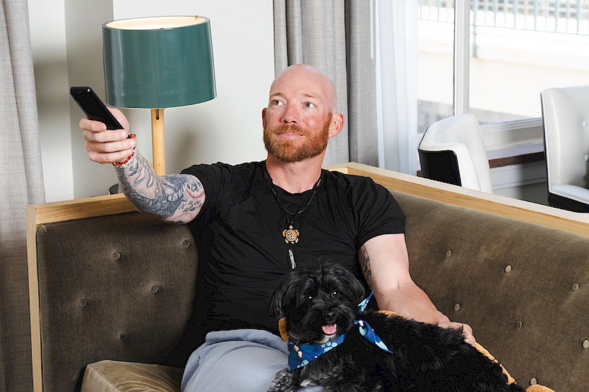 A man relaxes on a sofa with a dog, holding a remote. The room has a lamp and curtain, creating a cozy atmosphere.