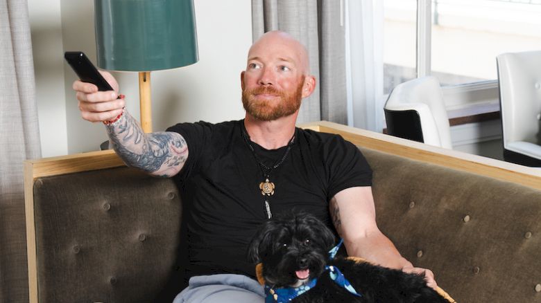 A man relaxes on a sofa with a dog, holding a remote. The room has a lamp and curtain, creating a cozy atmosphere.