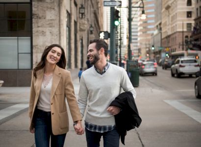 A couple is walking hand in hand on a city street, surrounded by buildings and cars, smiling and enjoying their time together.