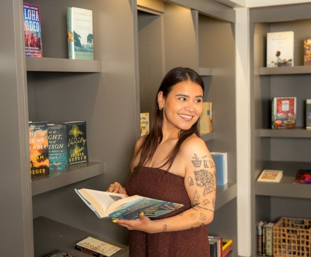 A person is standing in a library, holding an open book and looking at the shelves filled with various books.