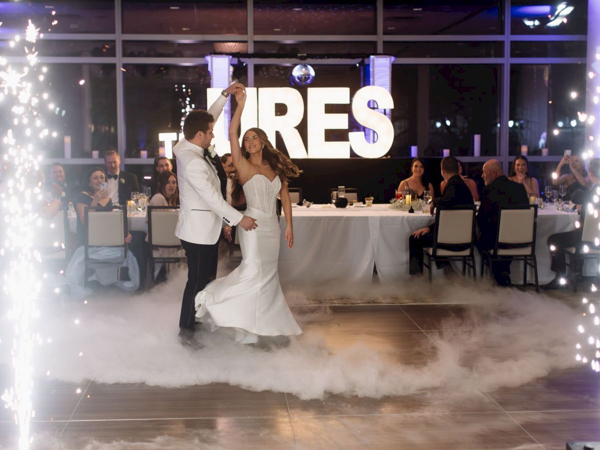 A couple dances on a fog-covered floor under fireworks, with a large illuminated sign in the background, surrounded by seated guests.