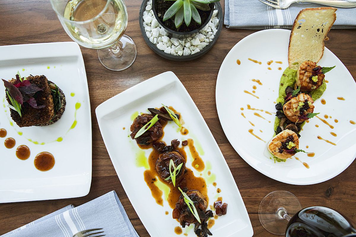 Three gourmet dishes beautifully plated on a wooden table, accompanied by wine glasses and a small plant centerpiece.