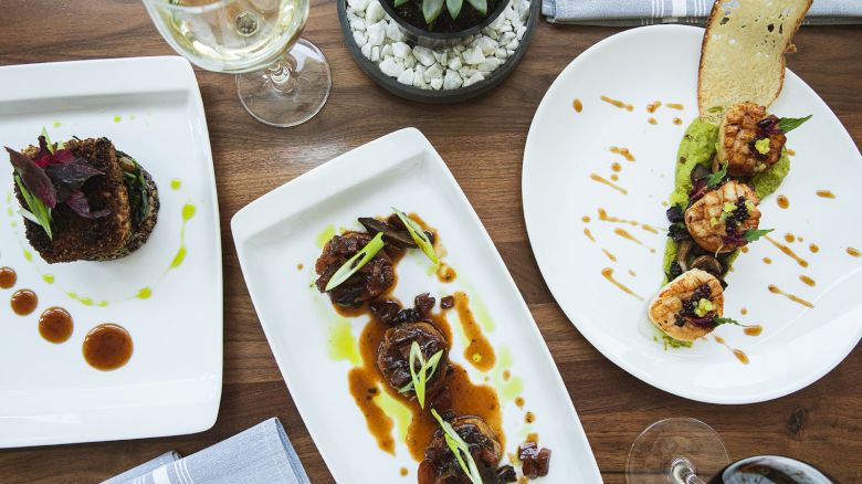 Three gourmet dishes beautifully plated on a wooden table, accompanied by wine glasses and a small plant centerpiece.