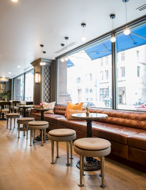 A cozy cafe interior with plush seating, round tables, and large windows letting in natural light from the street.