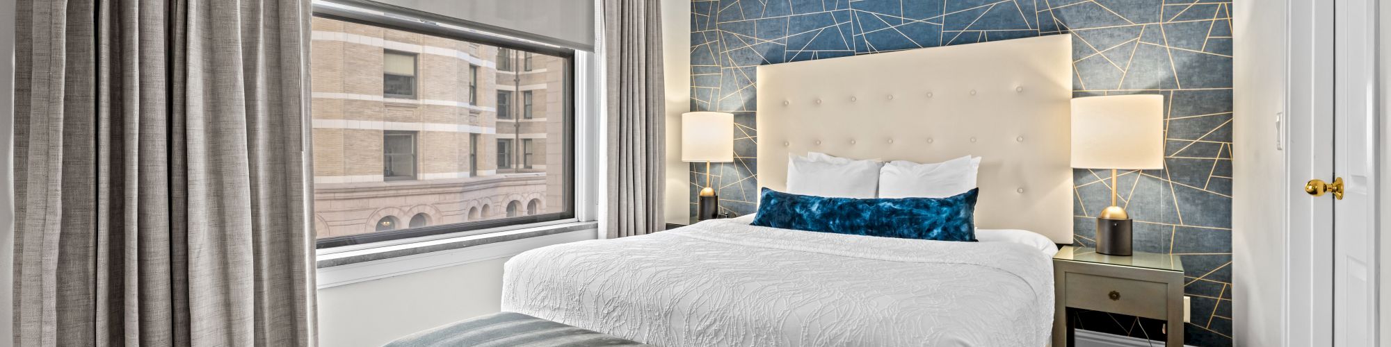 A modern bedroom with a bed, abstract wall design, side tables with lamps, a bench, and a window with curtains.