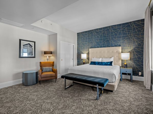 A modern bedroom with a king-size bed, geometric accent wall, armchair, side table, and bright window.