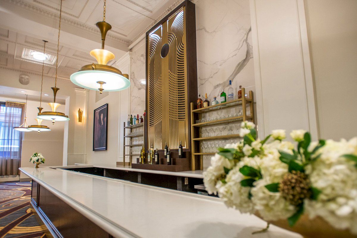 The image shows an elegant bar interior with decorative lighting, flower arrangements, and a stylish shelving unit with bottles.