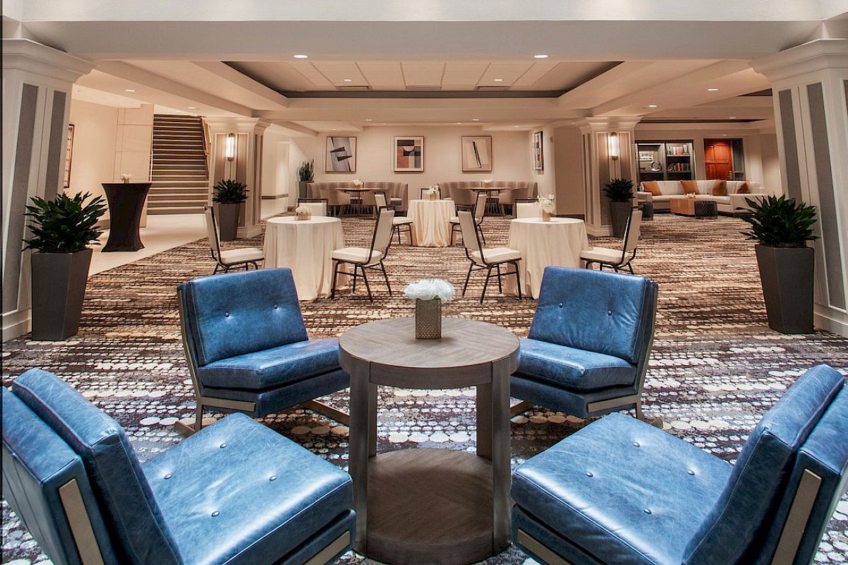 The image shows a modern hotel lobby with a glass ceiling, blue chairs around tables, and a bright, open space with plants.