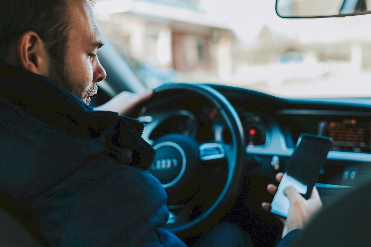 A person is sitting in a car, holding a smartphone while steering, with the car's dashboard and steering wheel visible.