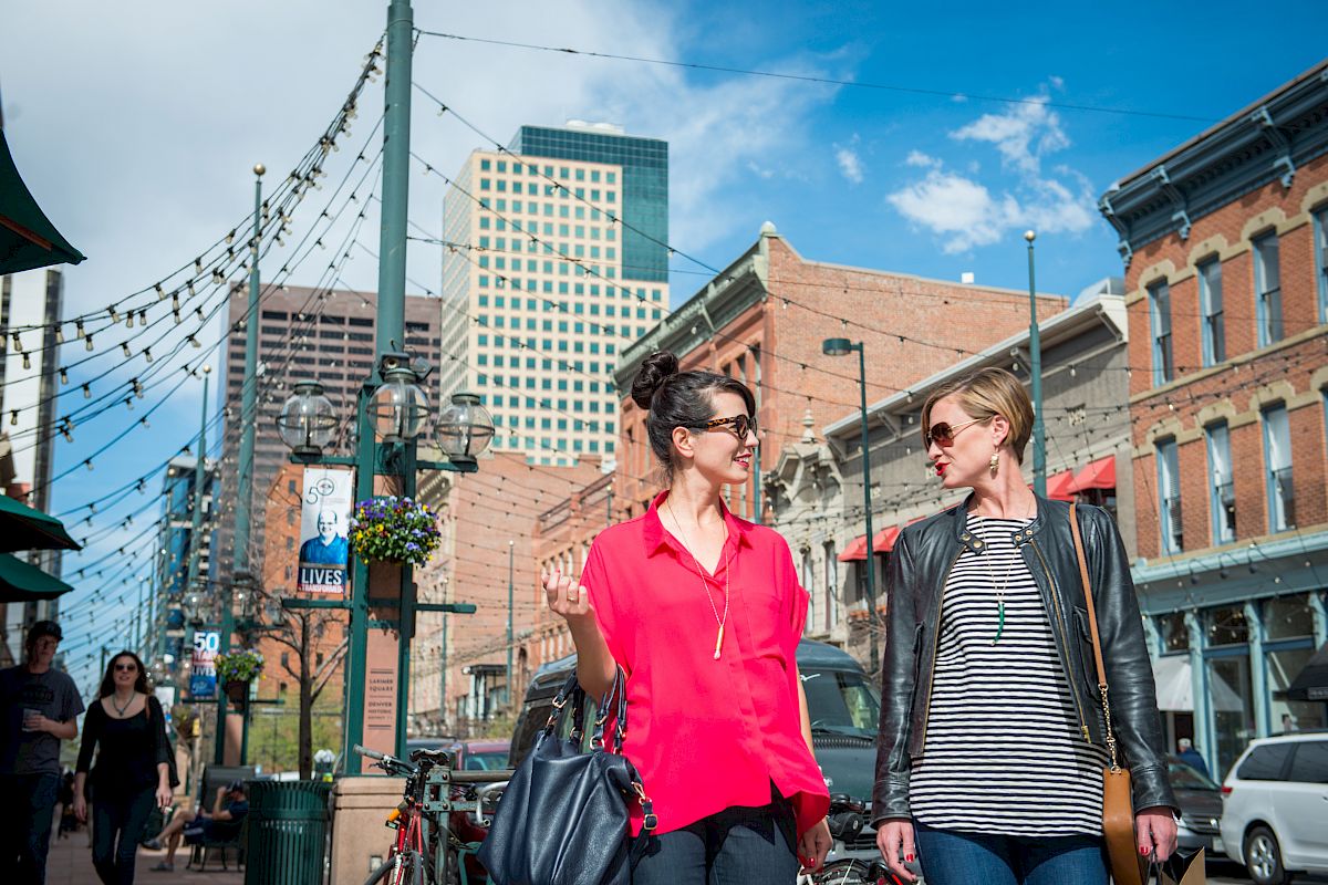 Two people walking and talking in an urban area with historical buildings, string lights, and blue sky in the background.