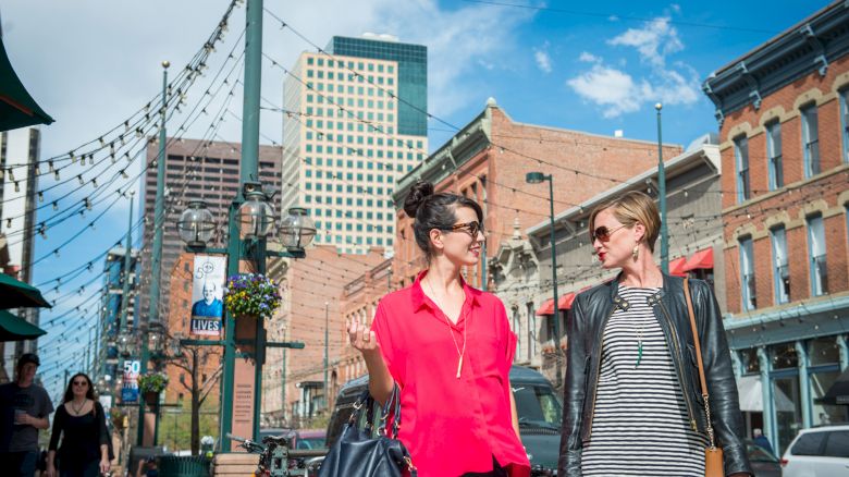 Two people walking and talking in an urban area with historical buildings, string lights, and blue sky in the background.