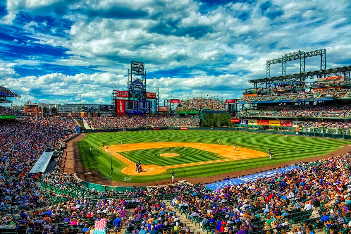 A packed baseball stadium under a vibrant blue sky hosts a game, with fans filling the stands and players on a bright, green field.