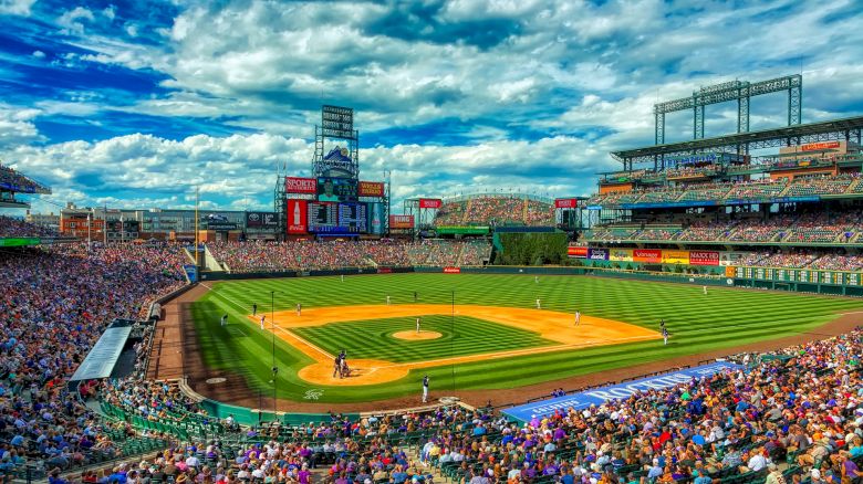 A packed baseball stadium under a vibrant blue sky hosts a game, with fans filling the stands and players on a bright, green field.