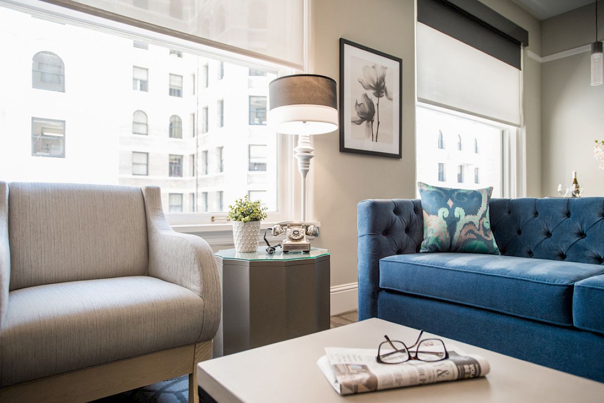 A cozy living room with a gray chair, blue sofa, lamp, framed art, and a table with glasses on a newspaper, near large windows.