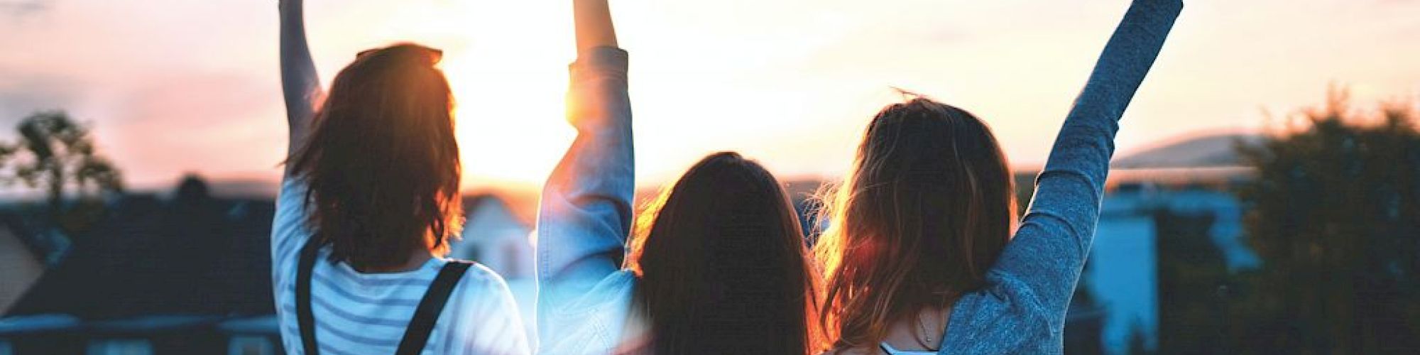 Three people stand on a rooftop at sunset, raising their hands in the air, with a scenic view and a warm, glowing sky in the background.