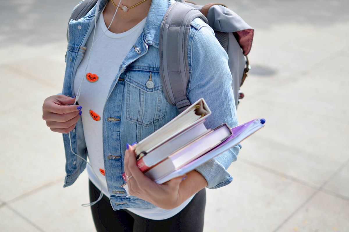 A person in a denim jacket carries books and a backpack, walking on a pavement.