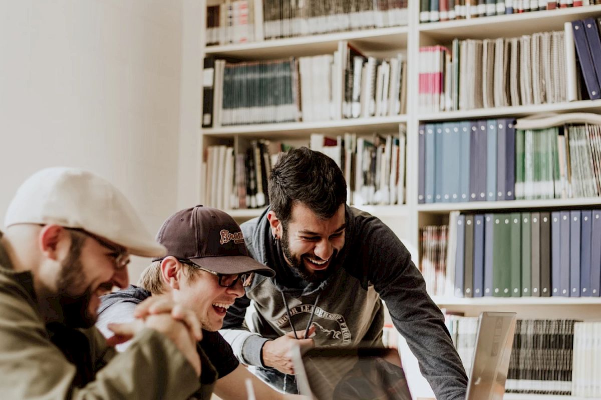 Three people are smiling and working at laptops in a library or office setting with bookshelves in the background.