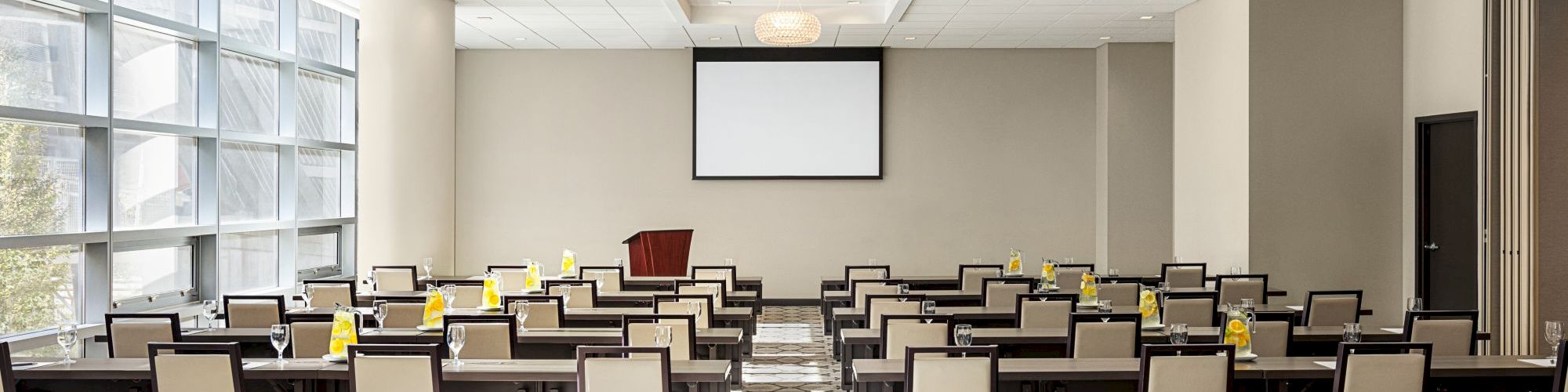 A conference room with rows of chairs and tables facing a screen, lit by natural light from large windows.