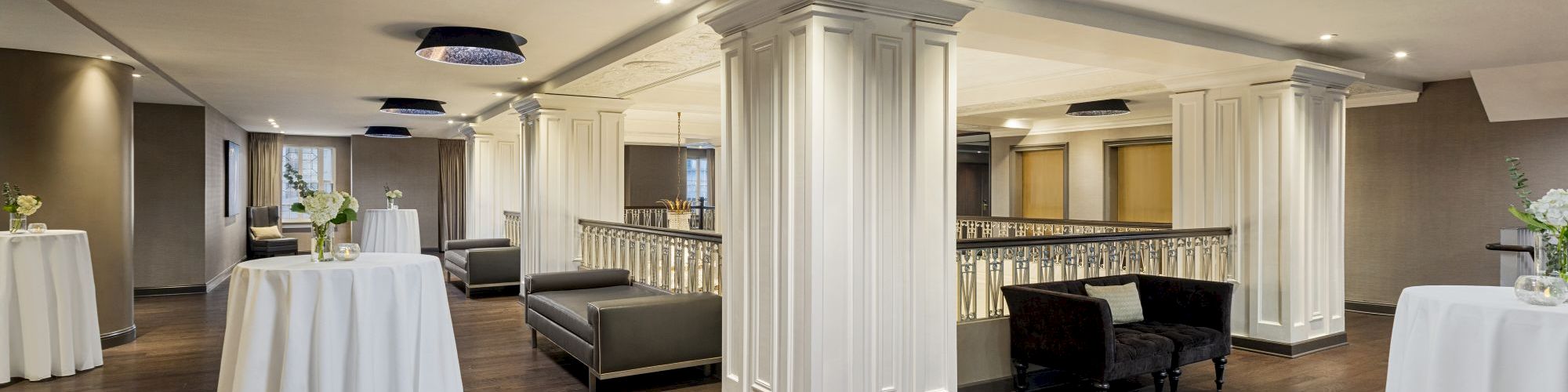 The image shows an elegant interior space with columns, cocktail tables, and seating, featuring a wooden floor and subdued lighting.