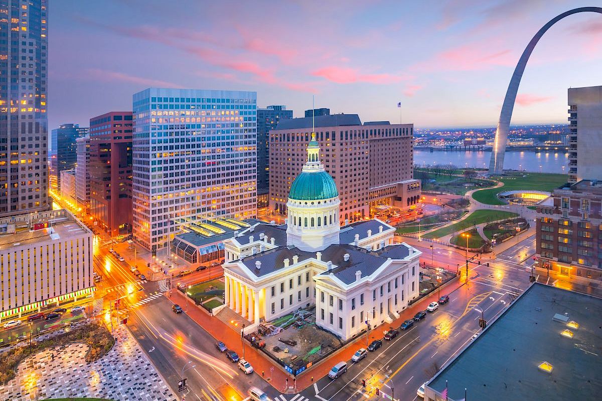 This image shows a cityscape with a historic domed building, surrounded by modern skyscrapers, and the Gateway Arch by the river at sunset.
