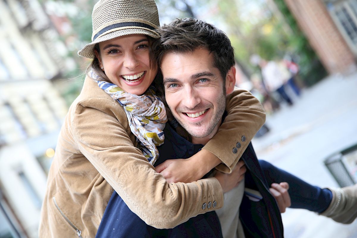 A smiling couple outside; the woman piggybacks on the man, both looking happy and casual in a relaxed, urban setting.