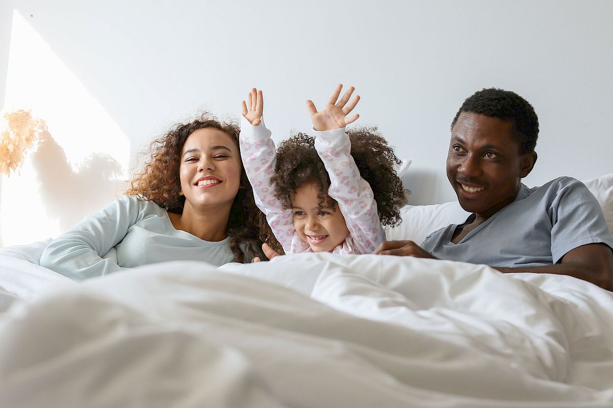 A family of three is lying in bed, smiling, with a child raising their arms joyfully between two adults.