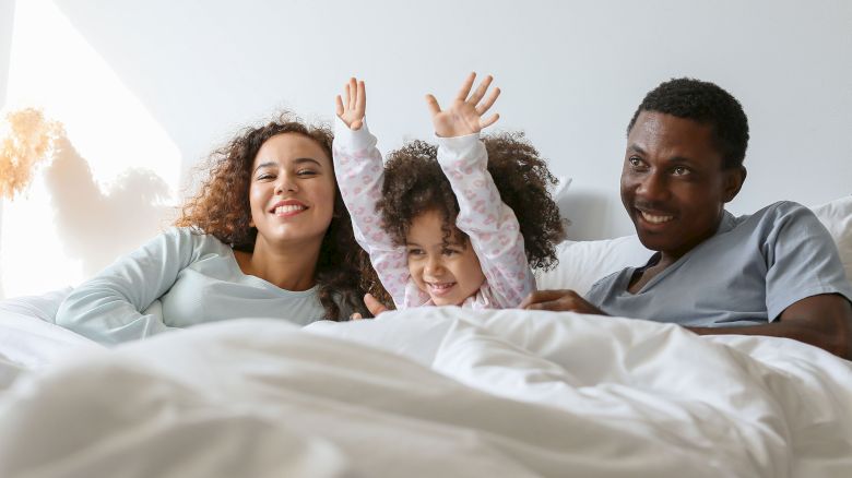 A family of three is lying in bed, smiling, with a child raising their arms joyfully between two adults.