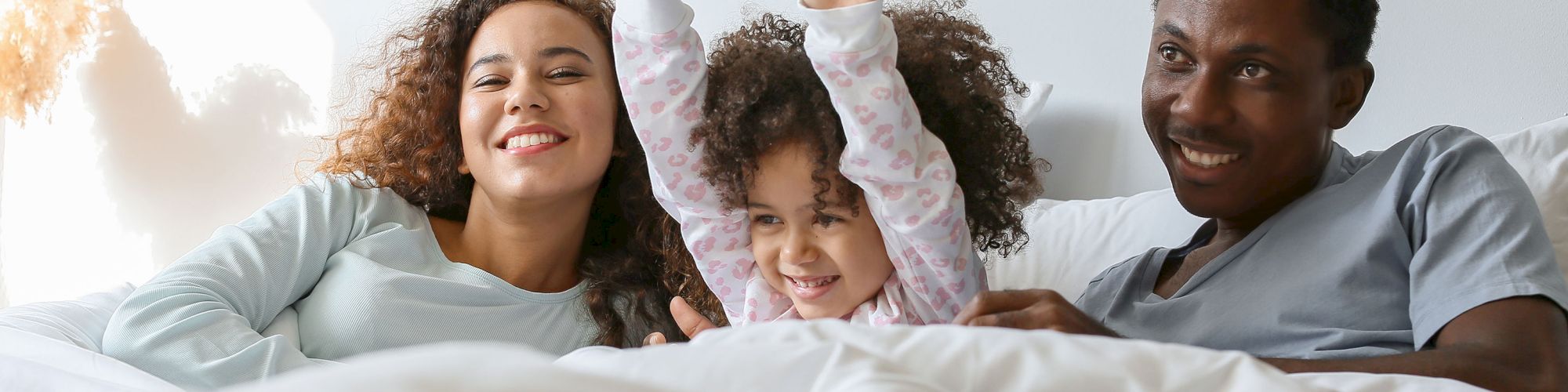A family of three is happily lying in bed together, with the child raising their arms in excitement.