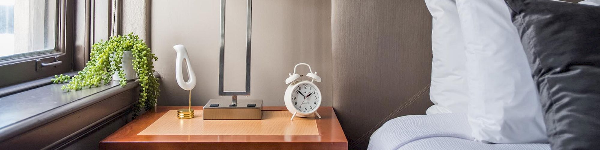 The image shows a neatly made bed next to a wooden nightstand with a lamp, a small plant, and a white alarm clock by a window.