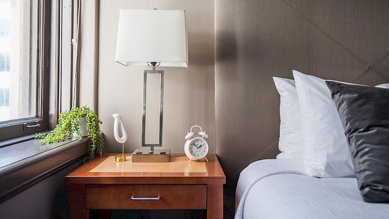 A bedside table with a lamp, alarm clock, and decorative items next to a bed with white and gray pillows.
