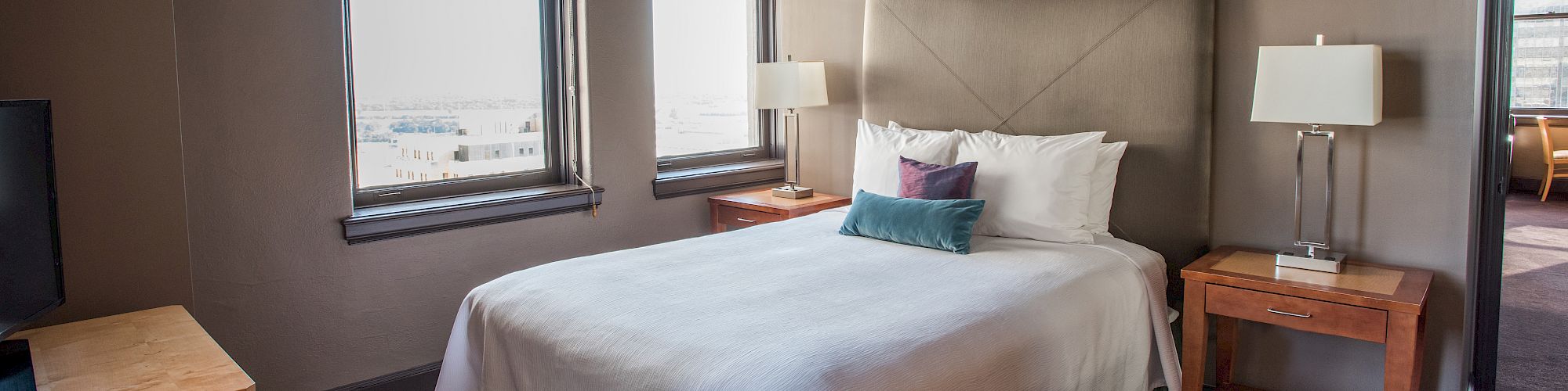 The image shows a neatly made bed in a hotel room with two windows, bedside tables, lamps, and a view outside.