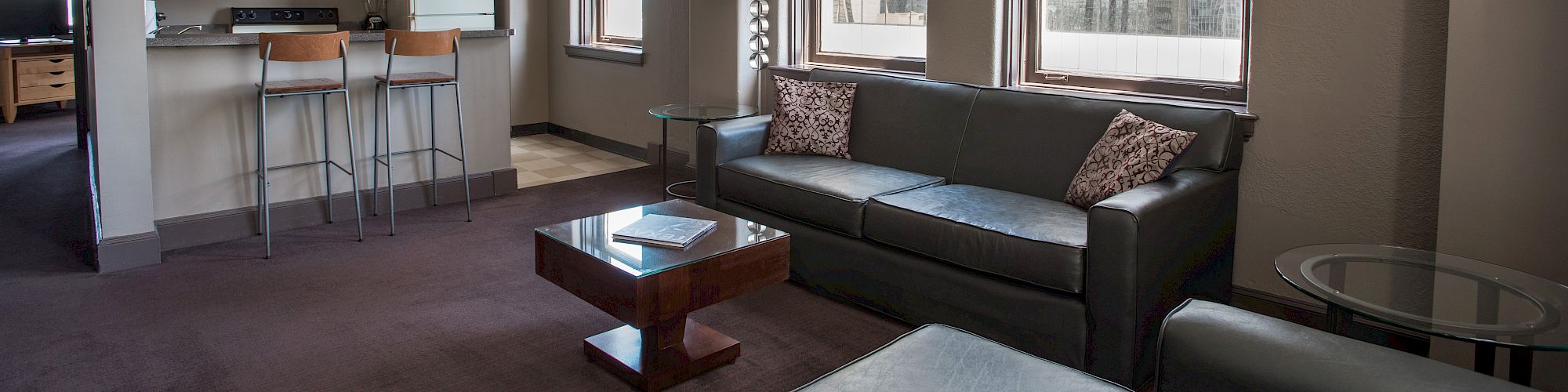 A modern living room with large windows, a gray sofa, and a kitchen area featuring wooden cabinets and bar stools.