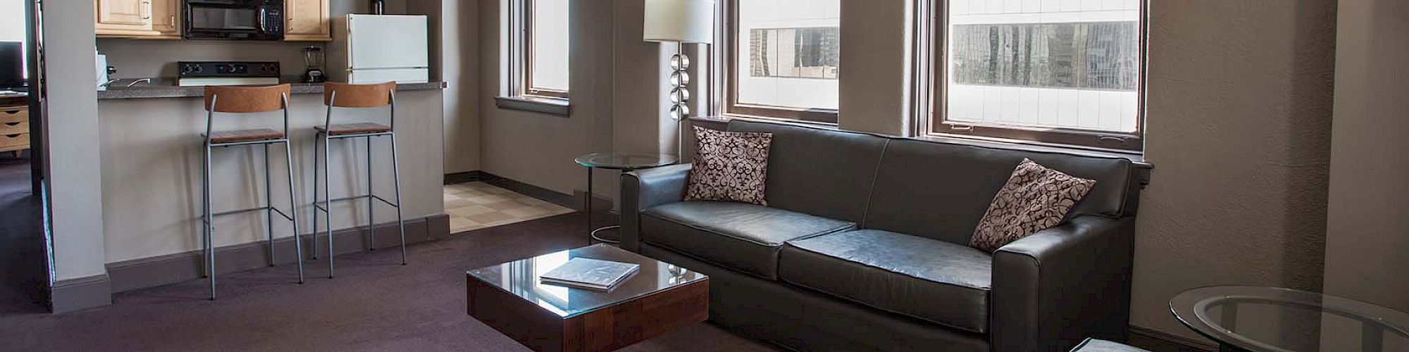 The image shows a modern living room with a sofa, lounge chair, coffee table, and a view into a kitchen area with barstools.