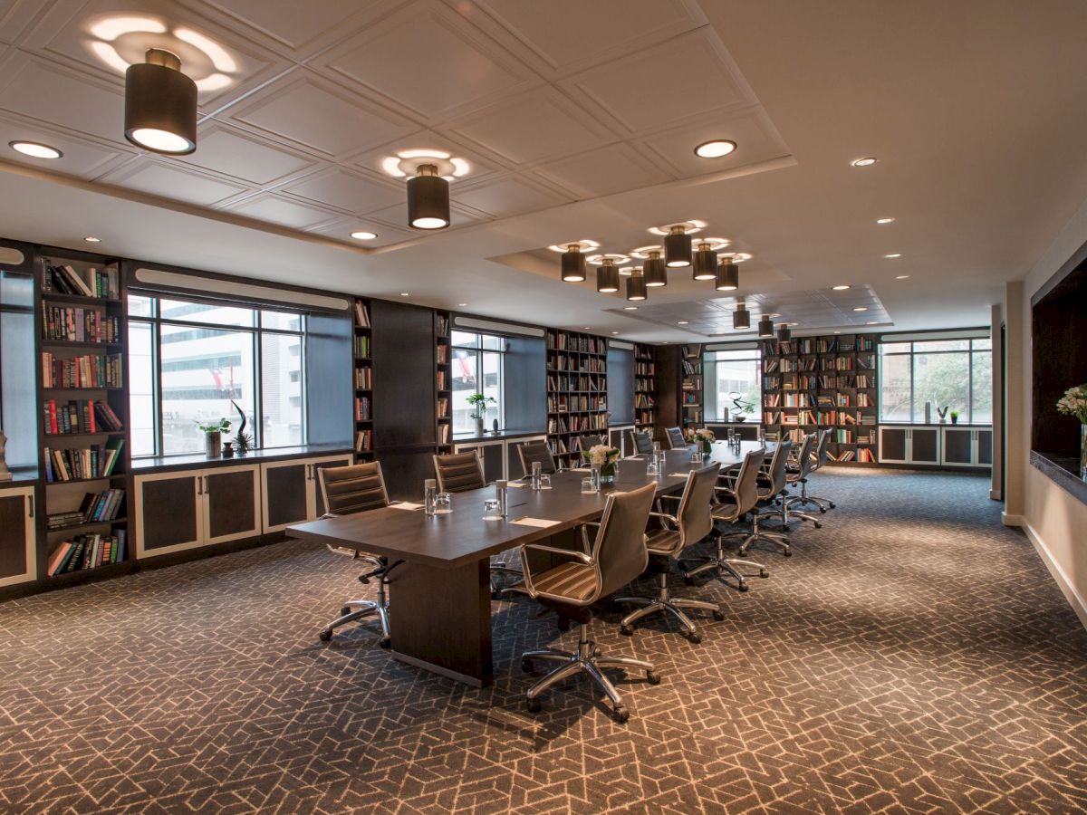 The image shows a modern meeting room with a long table, chairs, bookshelves, and large windows providing natural light.