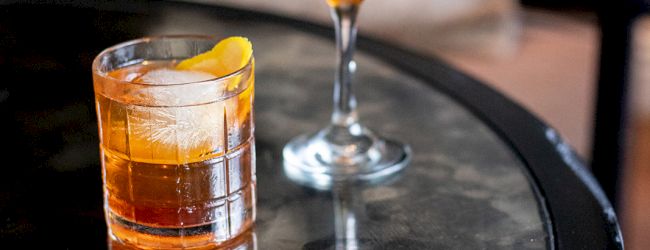 Two cocktails on a reflective table: one in a rocks glass with an orange peel, the other in a coupe glass with a cherry garnish.