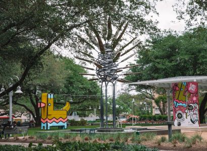 A park scene with colorful abstract sculptures, trees, and a pavilion in the background, surrounded by lush greenery.